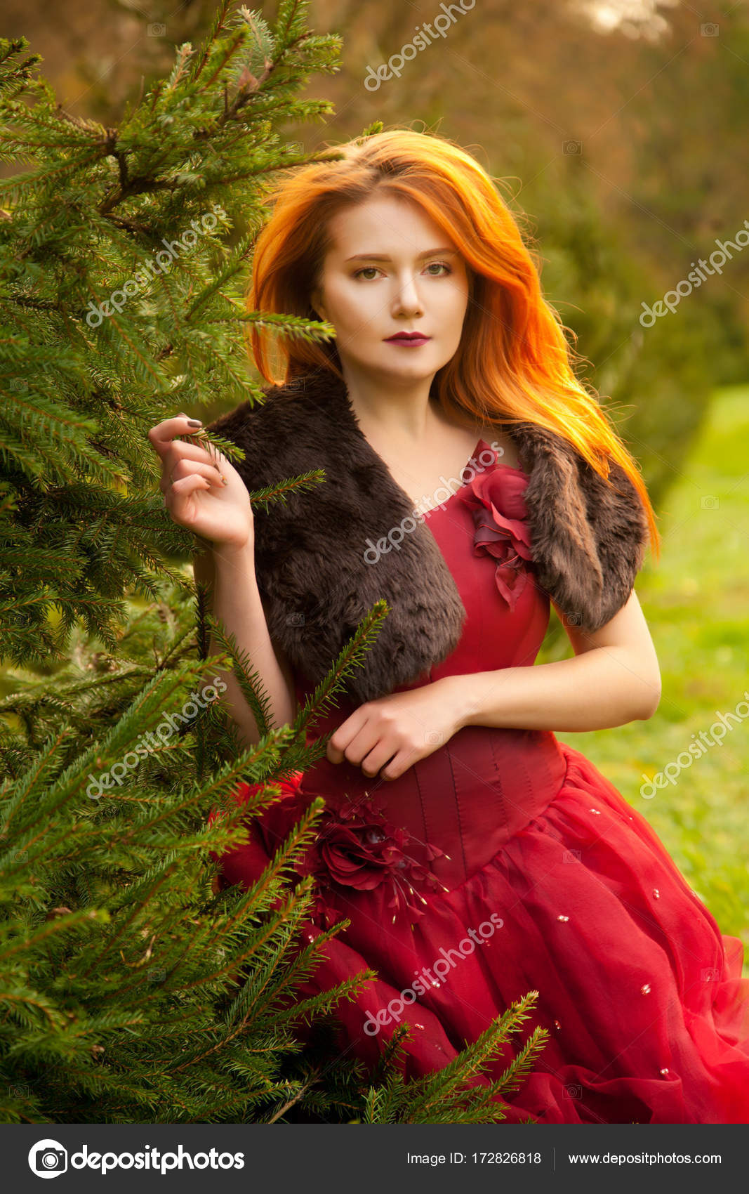 Beautiful young redhead girl wearing evening red dress walking in the fir tree alone Stock Photo by ©agnadevi