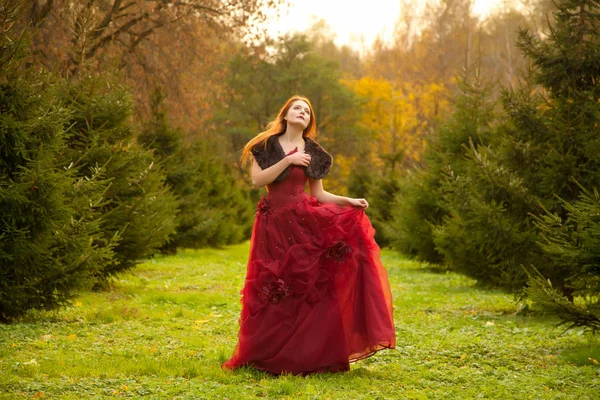 Красивая рыжая девушка, одетая в длинное вечернее красное платье и гуляющая в парке елки одна — стоковое фото