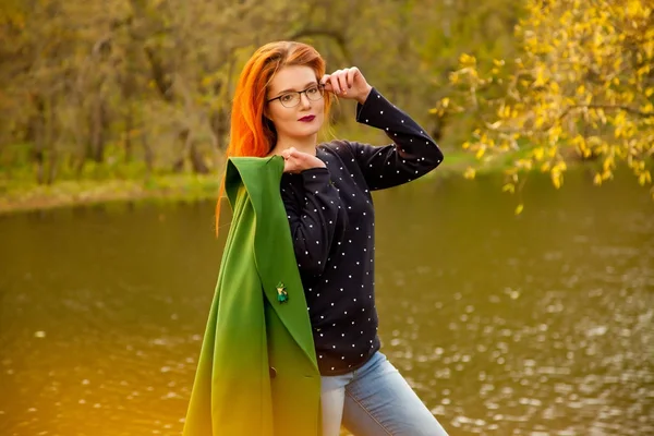 Bonita joven pelirroja vistiendo lindo suéter azul con pequeños corazones y sosteniendo su abrigo verde de pie cerca del frío lago de otoño en el parque de otoño — Foto de Stock