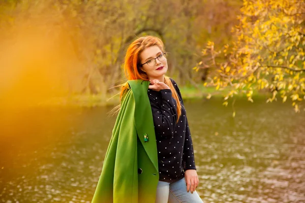 Csinos, fiatal vöröshajú nő viselt aranyos kék pulóver kis szíveket és a gazdaság zöld kabát állt az őszi parkban a hideg őszi-tónál — Stock Fotó