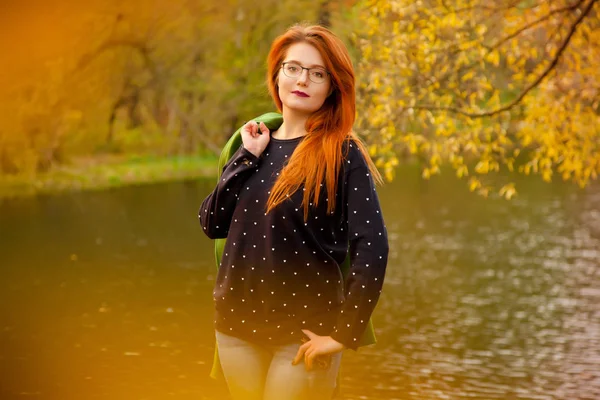 Pretty young redhead woman wearing cute blue sweater with little hearts and holding her green coat standing near the cold fall lake in the autumn park — Stock Photo, Image