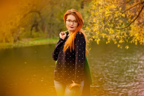 Bonita jovem ruiva mulher vestindo bonito camisola azul com pequenos corações e segurando seu casaco verde de pé perto do lago queda fria no parque de outono — Fotografia de Stock