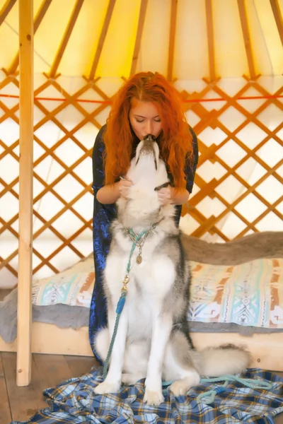 Bastante pelirroja mujer usando azul vestido de encaje sentado en la cama en su dormitorio y jugando con su mejor amigo Big Dog Malamute — Foto de Stock