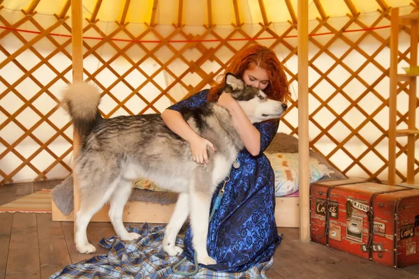 Mooie roodharige vrouw dragen van Blue Lace Dress zittend op het Bed In haar slaapkamer en het spelen met haar beste vriend grote hond Malamute — Stockfoto