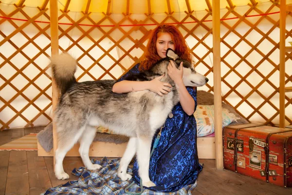Ładny Redhead dziewczyna ubrana w niebieski koronki sukni siedzi na łóżku w jej sypialni i bawi się z jej najlepszy przyjaciel duży pies Malamut — Zdjęcie stockowe
