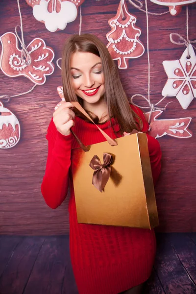 Pretty teenager girl wearing red knitted dress standing with golden shopping gift box and looking into the bag — Stock Photo, Image