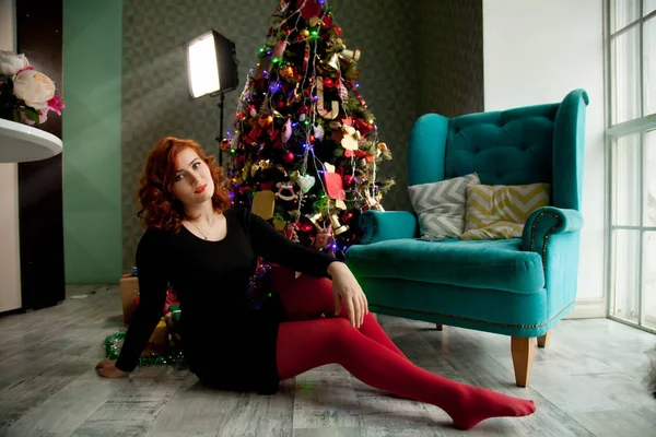 Bastante pelirroja chica sentada en el suelo cerca de la silla azul y el árbol de Navidad — Foto de Stock