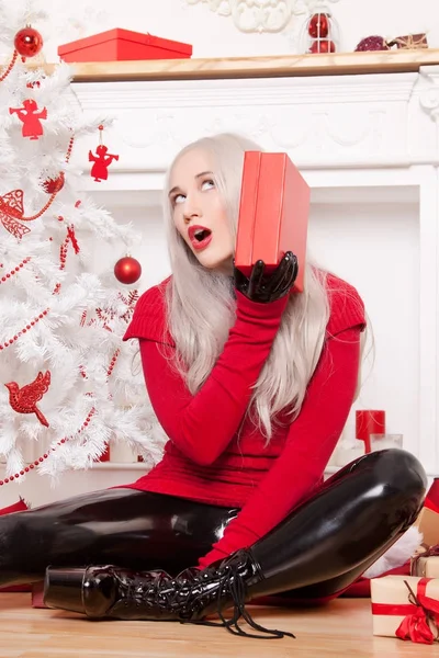 Hermosa mujer rubia de moda con traje de goma de látex negro, botas de tacón alto fetiche y suéter rojo Lond posando cerca del árbol de Navidad blanco y la chimenea con cajas de Año Nuevo de regalos —  Fotos de Stock