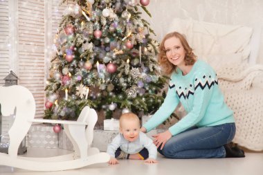 Bright giyen çok az anne kazak ve kot yeni yıl hediye kutuları ile dekore edilmiş Noel ağacı yanında küçük bebeği ile oynarken ördü. Gerçek aile.