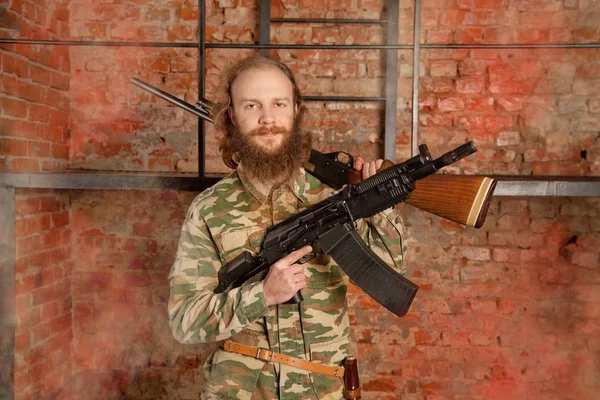 thin man with a beard wearing in military clothes and standing with a gun against a brick wall