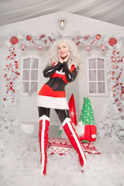 pretty blonde santa woman wearing red new year dress and shiny thigh high boots, standing near white winter snowy christmas decorated house alone