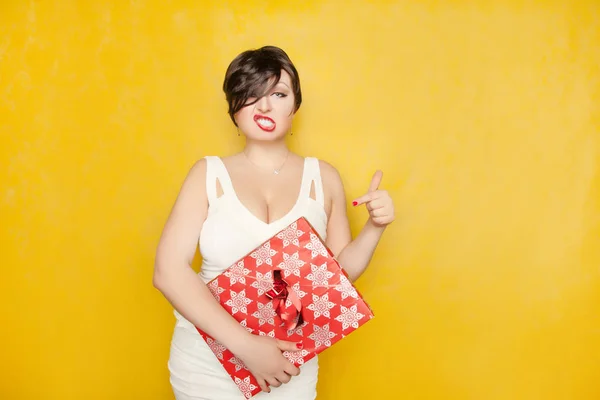 strict beautiful cruel girl brunette is in a white dress with a red gift in hand on yellow background. she is very unhappy and angry.