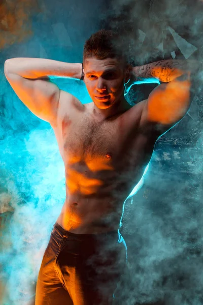 masculine young muscular male standing with naked torso in the smoke on a black background in Studio