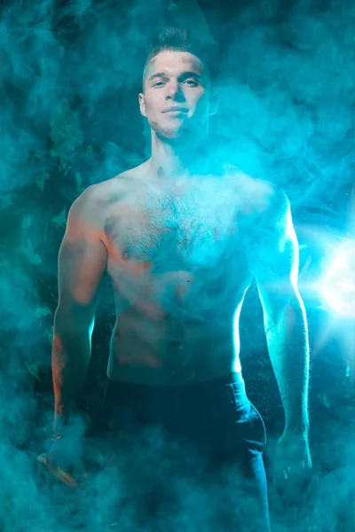 masculine young muscular male standing with naked torso in the smoke on a black background in Studio