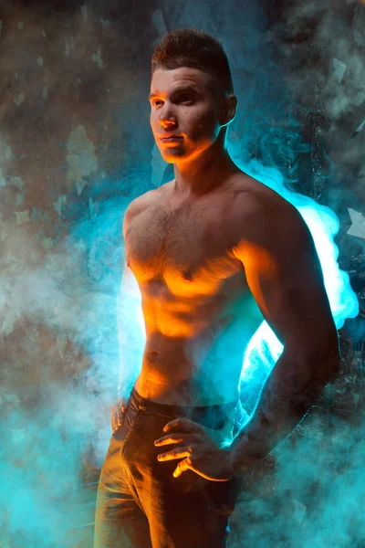 masculine young muscular male standing with naked torso in the smoke on a black background in Studio