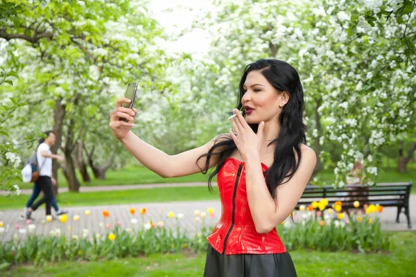 very bad and beautiful girl with black hair is dressed in a red corset, photographing herself doing selfie on a smartphone and smokes in the apple orchard among the flowers