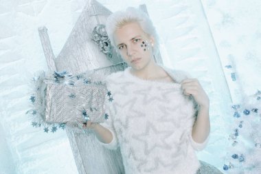 Beyaz süveterli, sarışın, beyaz tenli, yıldızlı sandalyede hediyesini tutan ve hediyesini açan adam.