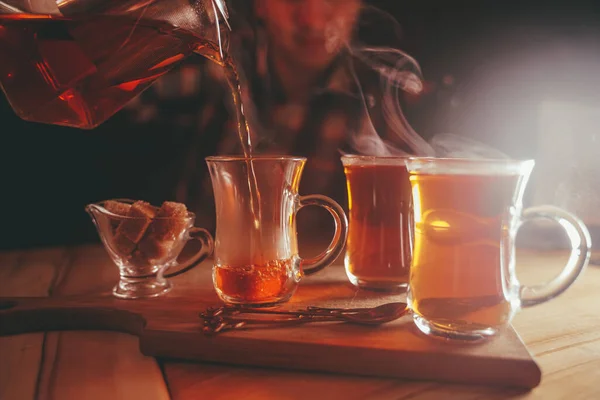 A young man pours tea from a hot teapot into a clear Cup on a wooden table in the evening on dark background — ストック写真