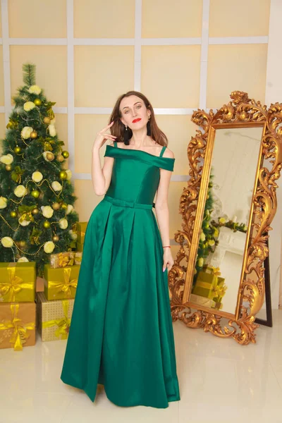 Belle femme mince gaie en robe longue soirée verte célébrer Noël dans une chambre blanche décorée près du grand miroir doré. corps pleine longueur — Photo