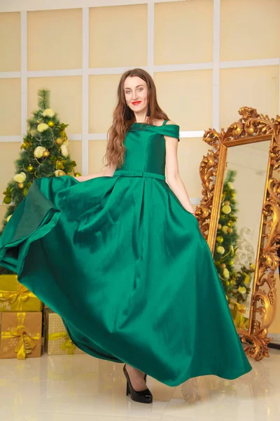 Schöne fröhliche schlanke Frau im langen grünen Abendkleid feiern Weihnachten in geschmückten weißen Raum in der Nähe des großen goldenen Spiegel. Ganzkörper — Stockfoto