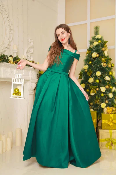 Красива весела струнка жінка в довгій зеленій вечірній сукні відзначає Різдво в білій прикрашеній кімнаті біля каміна — стокове фото