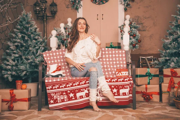 Affascinante ragazza sottile in jeans alla moda ricamato con perle e maglione di Natale a piedi nudi seduto su una panchina, corpo a figura intera — Foto Stock