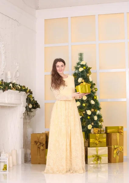 Beautiful slim caucasian adult woman in long evening yellow dress celebrates christmas indoors in her white room with decorated xmas tree — Stock Photo, Image