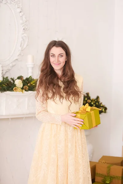 Красива струнка біла жінка в довгій вечірній жовтій сукні, що тримає подарункову коробку з бантом і святкує Різдво в приміщенні в її білій кімнаті біля каміна — стокове фото
