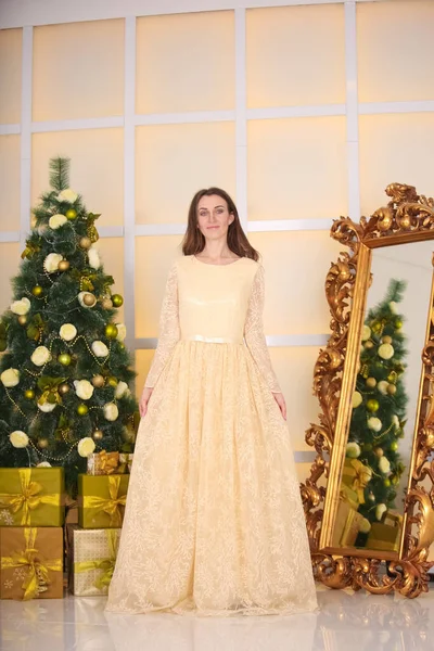 Beautiful slim caucasian adult woman in long evening yellow dress celebrates christmas indoors in her white room near the decorated big xmas tree — ストック写真