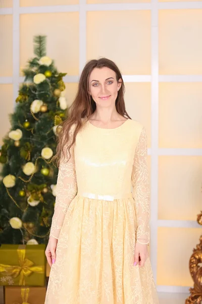 Красива струнка біла жінка в довгій вечірній жовтій сукні відзначає Різдво в приміщенні в її білій кімнаті біля прикрашеної великої ялинки — стокове фото