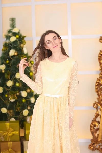 Bella donna adulta caucasica sottile in abito lungo da sera giallo celebra il Natale al chiuso nella sua stanza bianca vicino al grande albero di Natale decorato — Foto Stock