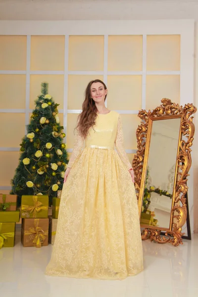Beautiful slim caucasian adult woman in long evening yellow dress celebrates christmas indoors in her white room near the decorated big xmas tree — Stock Photo, Image