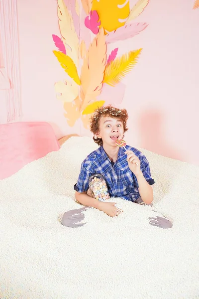 Adolescente chico en la piscina con espuma suave pequeñas bolas blancas. persona dedicada a la terapia de relajación. niño caucásico con piruleta de colores en el palo sobre fondo rosa . — Foto de Stock