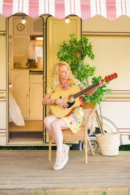 Uzun sarı saçlı, sevgi dolu orta yaşlı kız arkadaş karavanın yanında gitar çalıyor ve şarkı söylüyor.