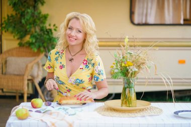 Sarı elbiseli, çekici, mutlu, sarışın kız karavan kampı minibüsünün arka planında bir masada oturuyor.