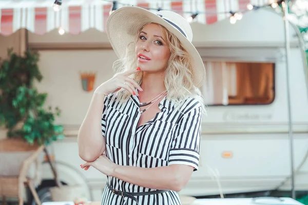 a cute woman with wavy blonde hair in a trendy summer striped dress on the background of a trailer camper van rests