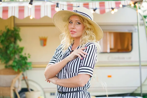 a cute woman with wavy blonde hair in a trendy summer striped dress on the background of a trailer camper van rests