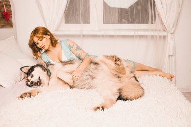 Büyük köpeğiyle poz veren gecelikli, kızıl saçlı, beyaz yatakta en iyi arkadaşı Malamute olan çekici ve uzun boylu bir kız.