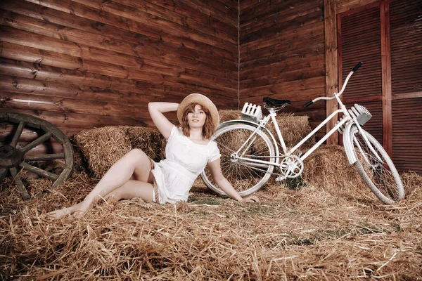 Mooi meisje in een witte zomer jurk poseert met een fiets in het hooi na de herfst oogst — Stockfoto