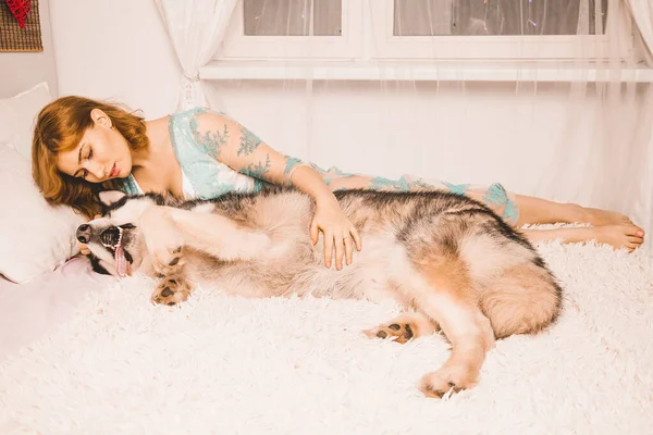 Чарівна і руда дівчина з рудим волоссям у нічній сукні зі своїм великим собакою, найкращим другом Маламута в білому ліжку в спальні. — стокове фото
