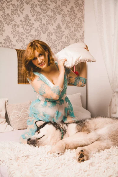 Encantadora chica de tamaño grande con el pelo rojo en un camisón posando con su perro grande, un mejor amigo Malamute en la cama blanca en el dormitorio — Foto de Stock