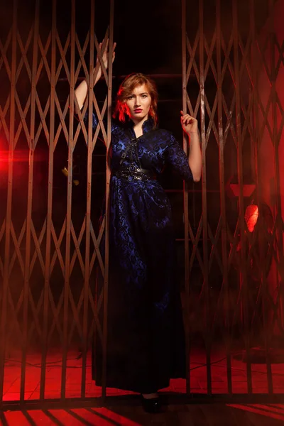 The red haired plus size woman in evening long dress and leather harness standing near big gates in a black alley in night — Stock Photo, Image