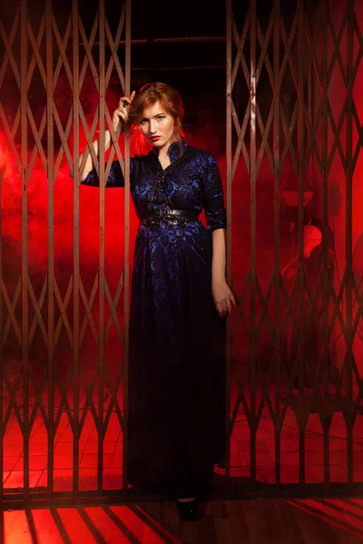 Rudowłosy plus rozmiar kobieta w wieczorowej długiej sukience i skórzanej uprzęży stojącej w pobliżu dużych bram w czarnej alejce w nocy — Zdjęcie stockowe