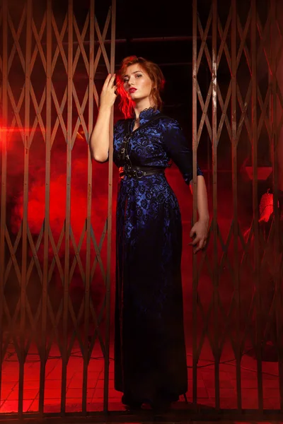O cabelo vermelho plus tamanho mulher à noite vestido longo e chicote de couro em pé perto de grandes portões em um beco preto à noite — Fotografia de Stock