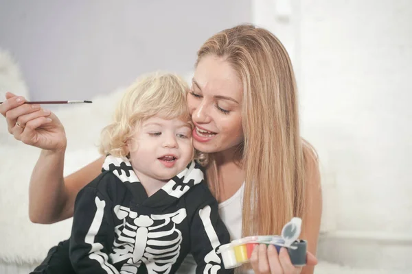 Young woman and her baby son have autumn fun with pumpkin on white windowsill, boy dressed in skeleton costume — Stock Photo, Image