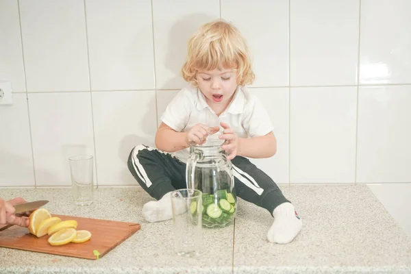Πορτρέτο μιας αξιολάτρευτης μητέρας και του μικρού παιδιού προετοιμασία ένα ποτό φυτικό νερό μαζί στη λευκή κουζίνα — Φωτογραφία Αρχείου