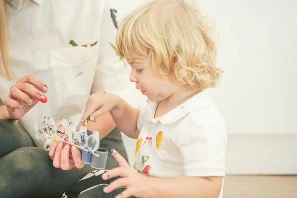 A cheerful mother and a joyful child sit on the floor in white t-shirts and paint each other with paints — Stock Photo, Image