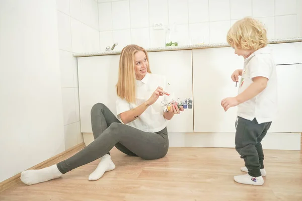 A cheerful mother and a joyful child sit on the floor in white t-shirts and paint each other with paints — Stock Photo, Image