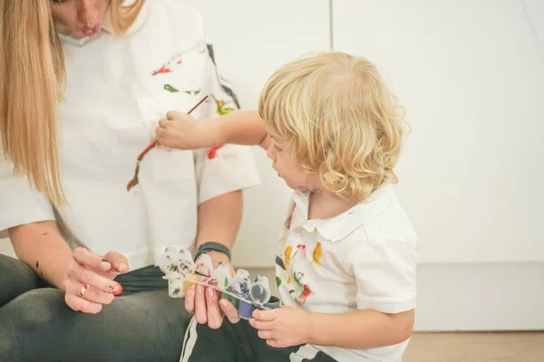 A cheerful mother and a joyful child sit on the floor in white t-shirts and paint each other with paints — Stock Photo, Image