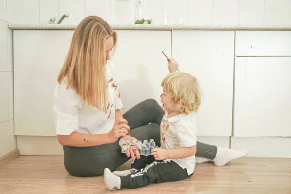 A cheerful mother and a joyful child sit on the floor in white t-shirts and paint each other with paints — Stock Photo, Image